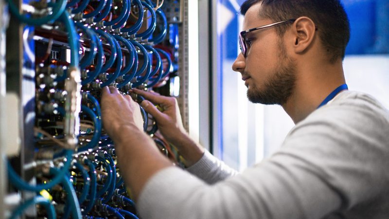Side  view portrait of young man connecting cables in server cabinet while working with supercomputer in data center