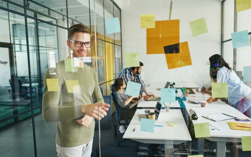 Amicable young man in glasses entering the office and pointing to a sticky note on the wall
